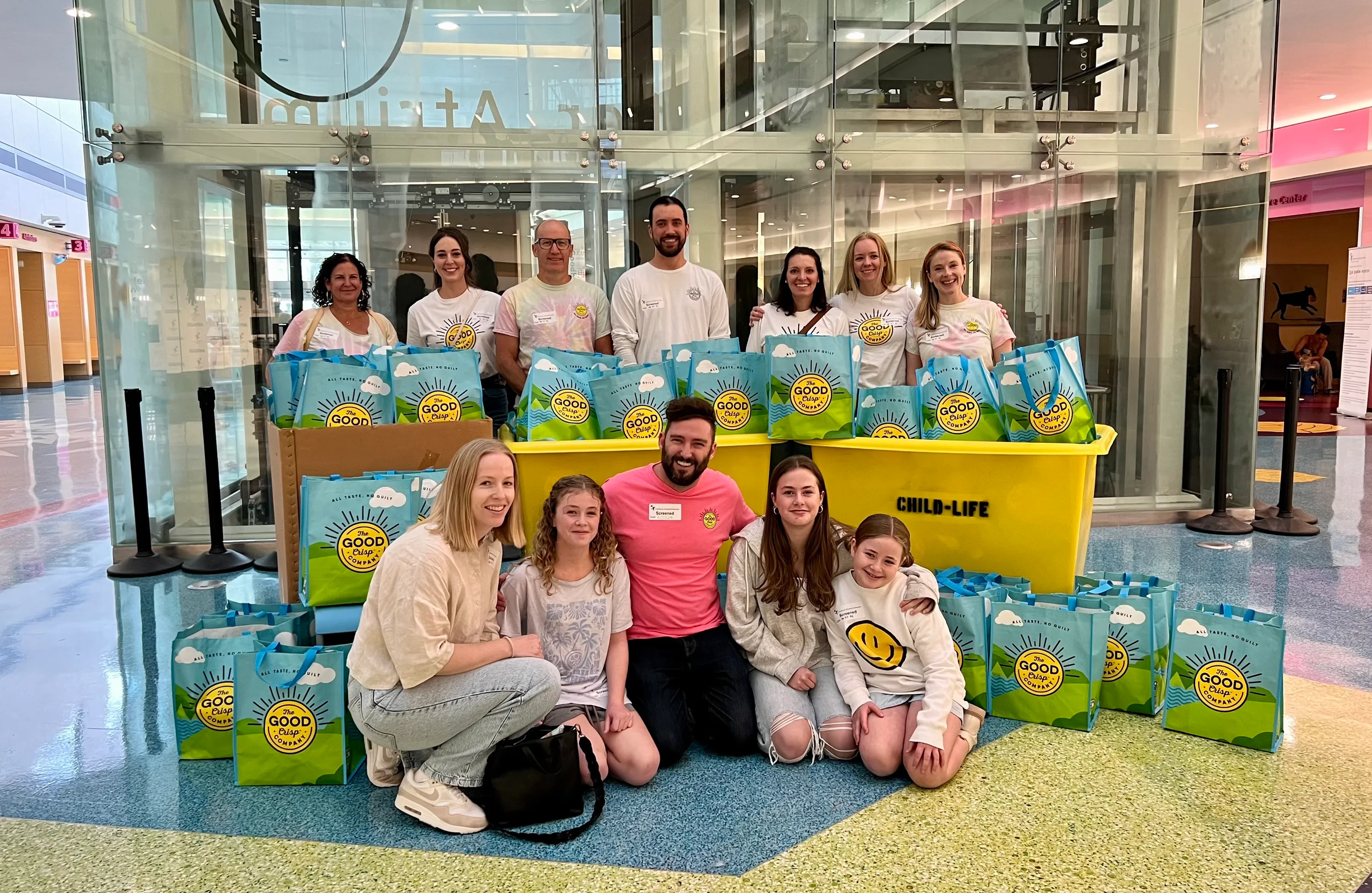 Volunteers and families gathered in a hospital atrium around yellow Child-Life donation bins filled with Good Crisp gift bags