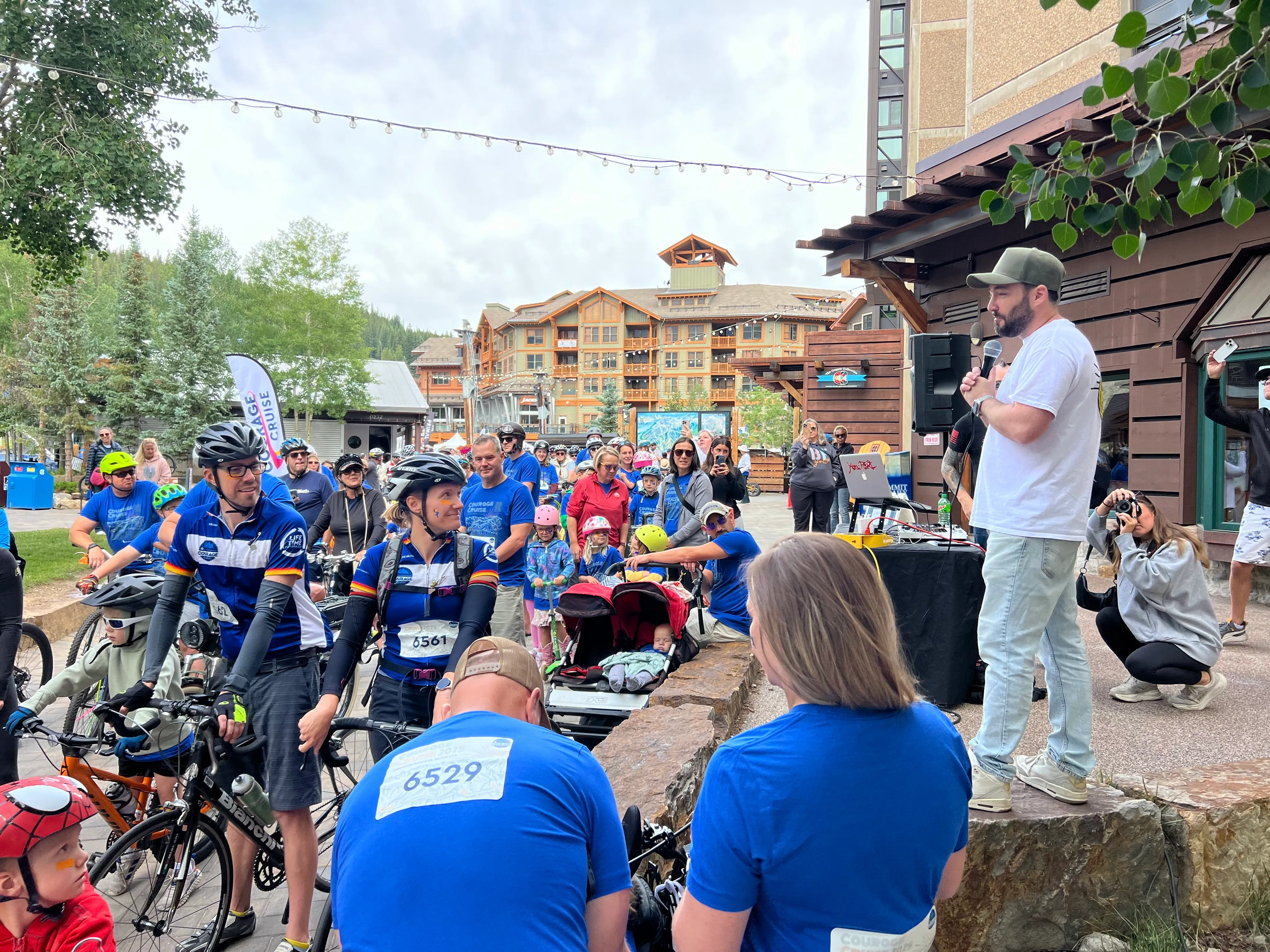 Crowd of cyclists in helmets and jerseys listening to a speaker at an outdoor charity ride event