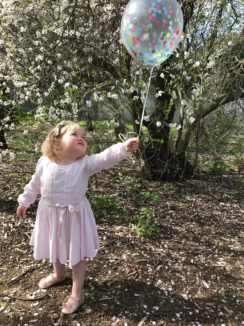 Trixie's sister in a white dress, holding a confetti balloon under a blossom tree
